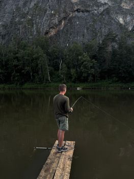 Adult man fishing from a wooden dock by a lake with a forest and rocky cliff in the background.