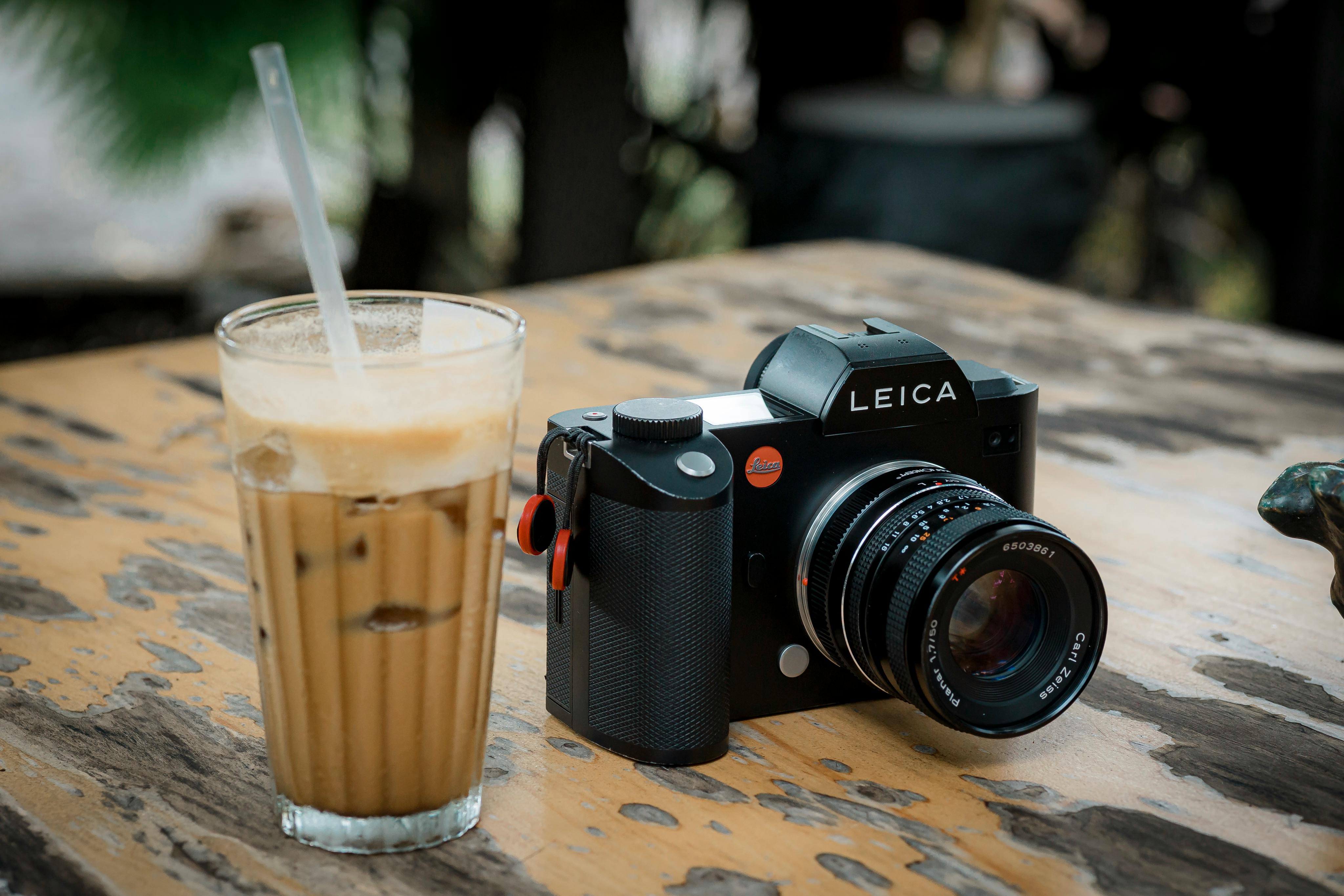 A Leica camera and a glass of iced coffee on a rustic wooden table outdoors.