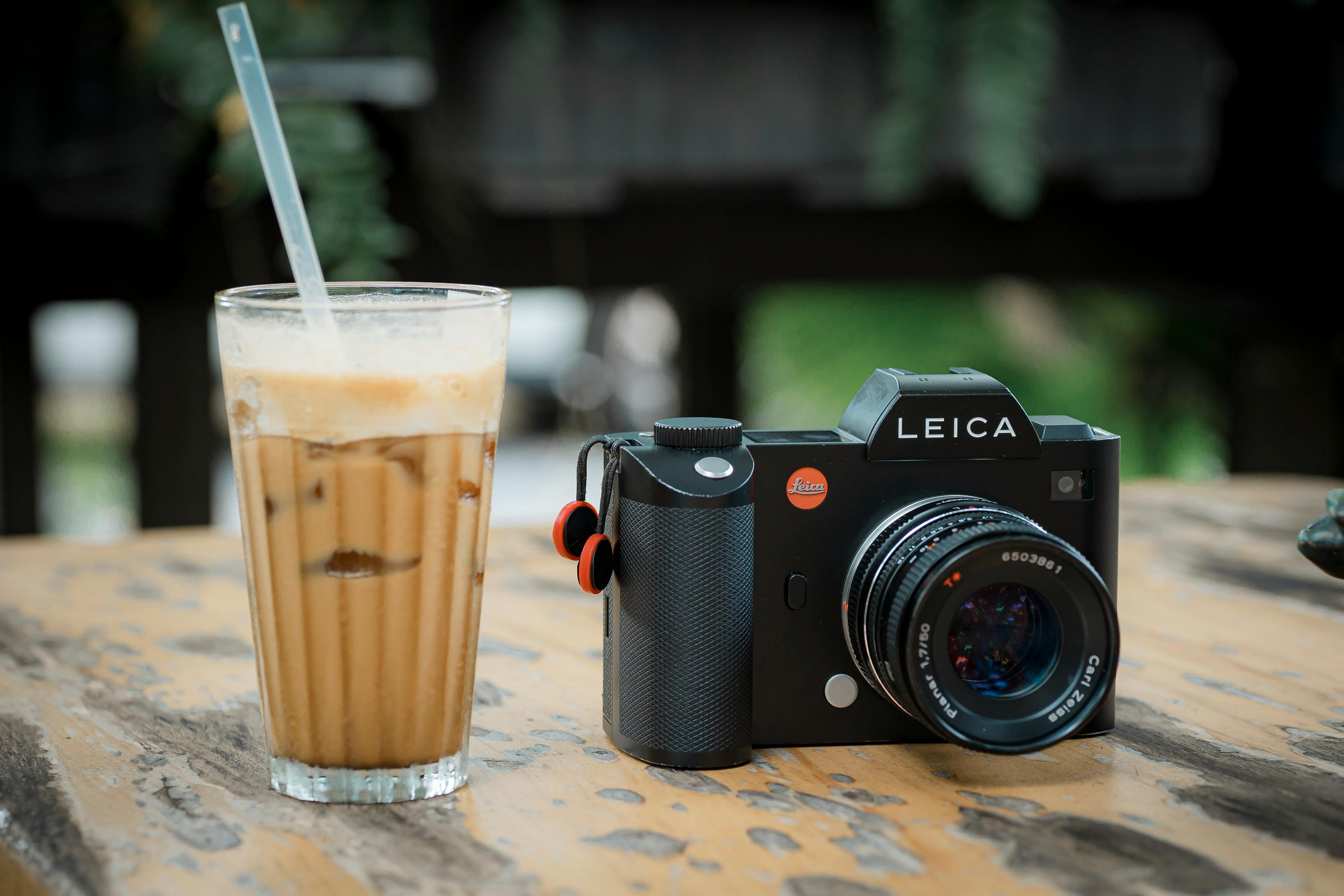A stylish camera sits next to an iced coffee on a rustic wooden table outdoors.