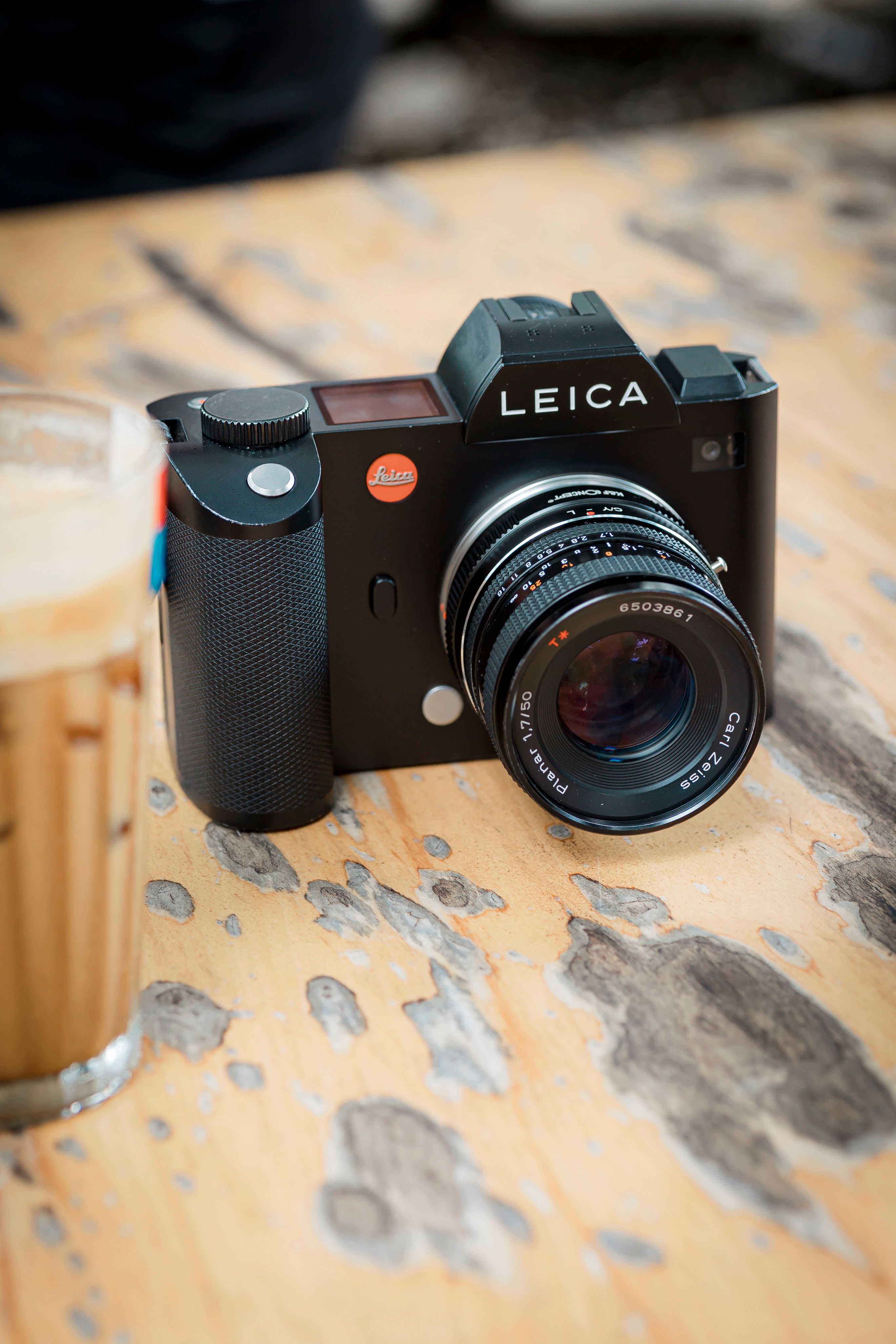 Close-up of a Leica camera on a weathered wood table beside a cold coffee.