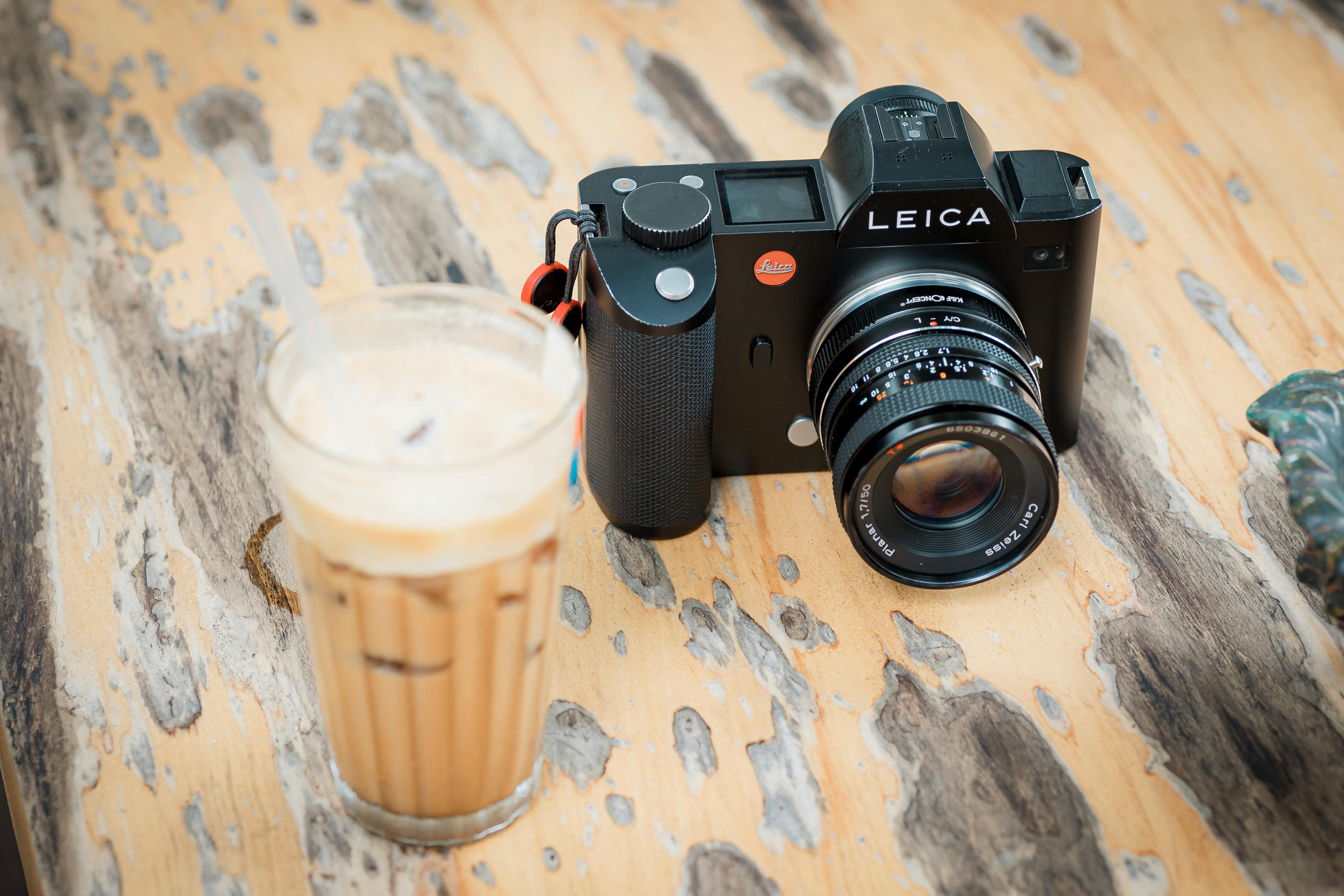 Leica camera with iced coffee on rustic wooden table, outdoors.