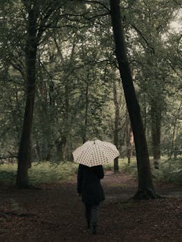 A solitary woman walks through a lush forest holding a white umbrella on a rainy day.