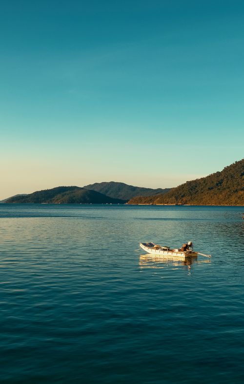 Free A serene boat floats on calm water against a backdrop of hills under a clear blue sky at sunset. Stock Photo