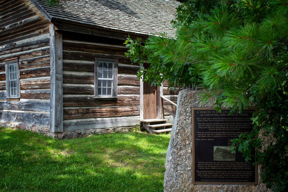 A rustic log cabin with a historical plaque surrounded by green foliage.
