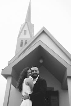 Black and white photo of a happy couple embracing, standing before a church.