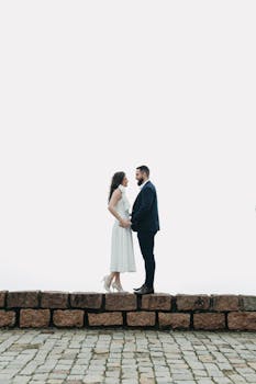 Romantic couple in elegant attire standing on a stone wall against a clear sky.