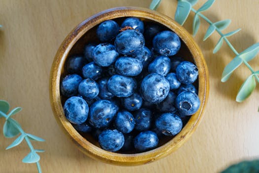 A top view of fresh blueberries in a wooden bowl, surrounded by green leaves, on a wooden table.