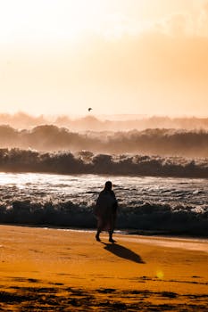 A solitary figure walks along a serene beach at sunset, creating a peaceful, reflective scene.