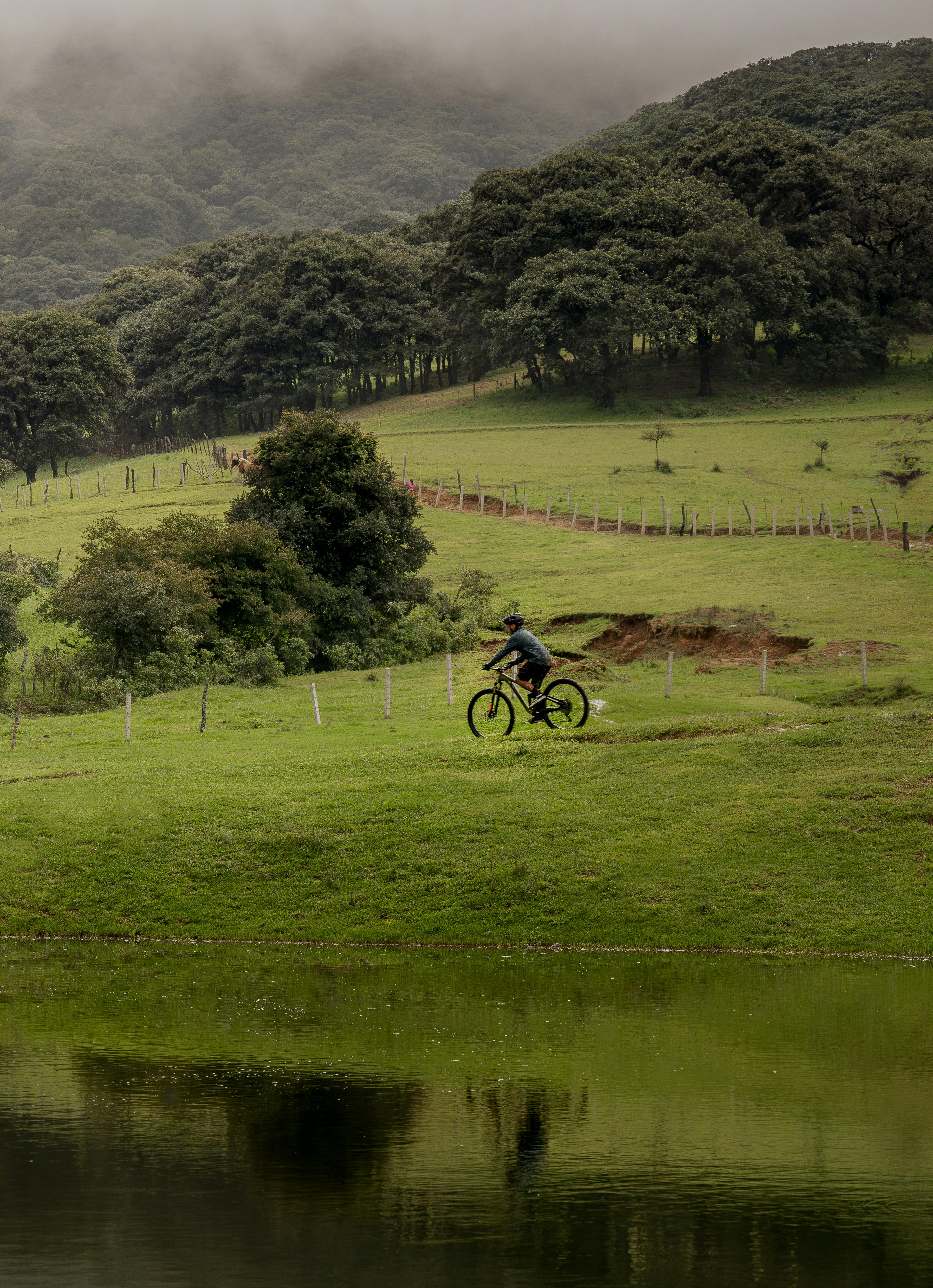 Cyclist riding through scenic San Luis Potosí landscape with lush hills and tranquil lake reflection.