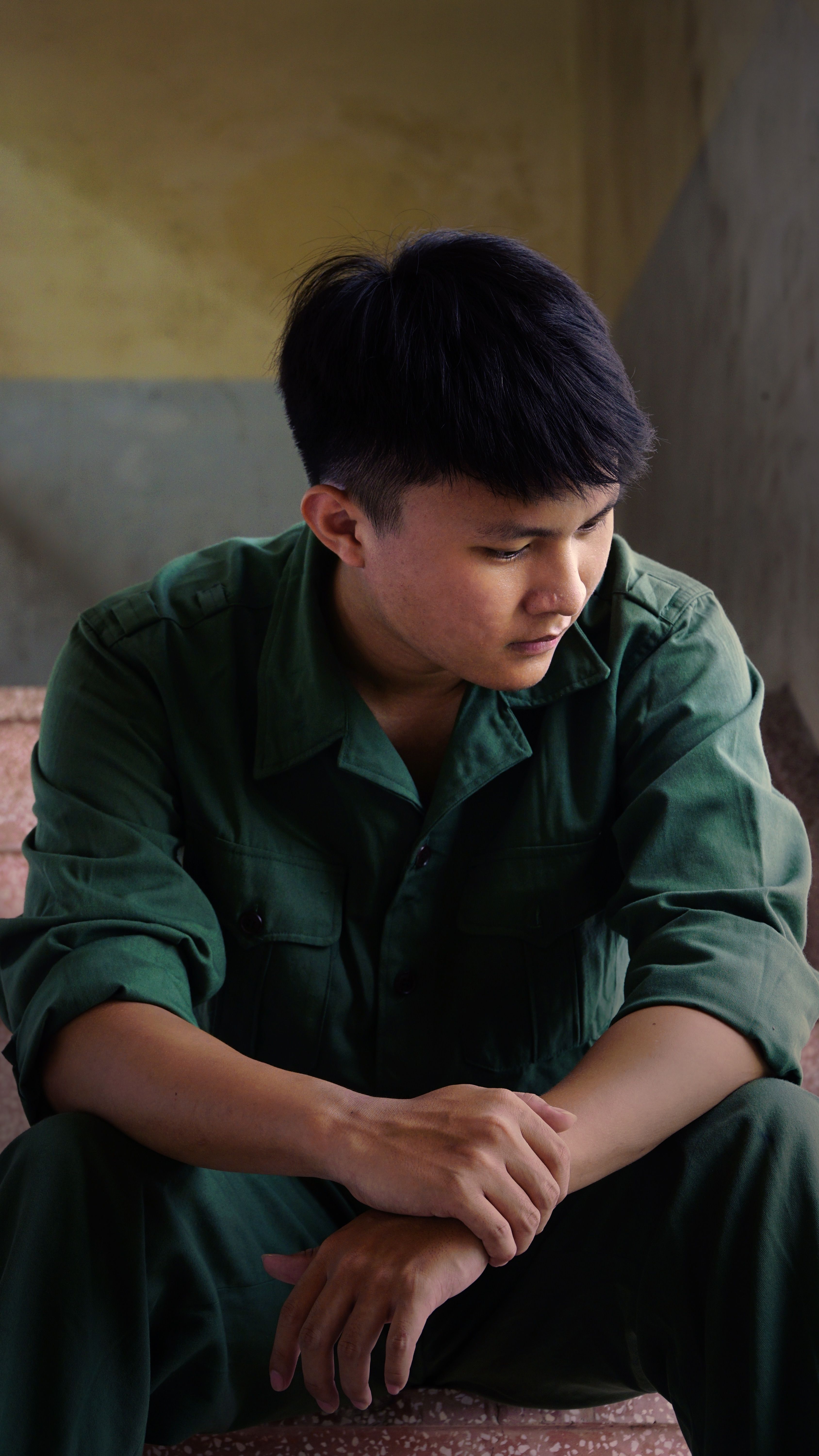 Young man in green overalls sitting indoors, looking contemplative in soft lighting.