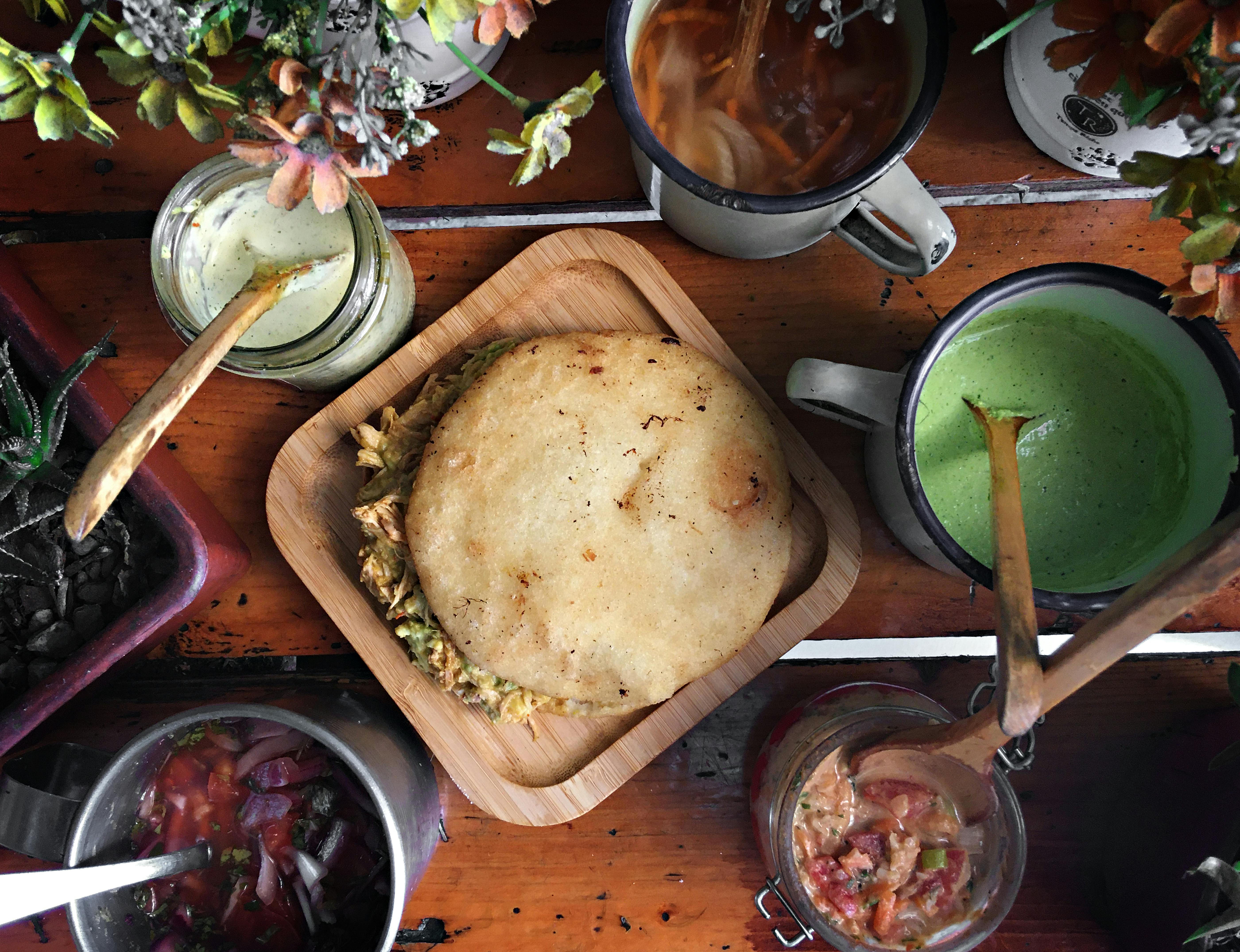Top view of a traditional Venezuelan arepa with assorted sauces and drinks on a rustic table.