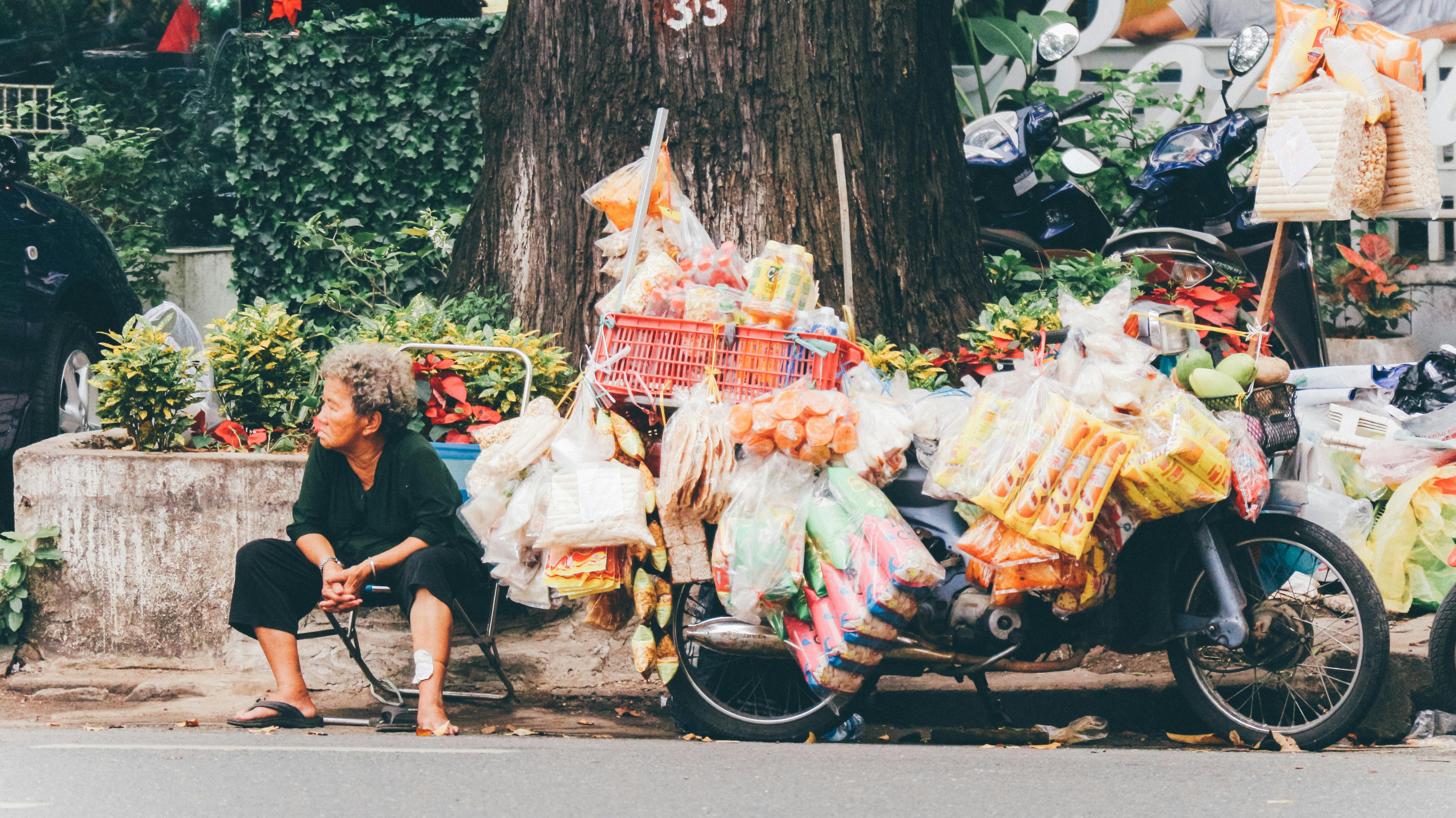 An elderly woman sits beside a street stall full of colorful snacks on a motorcycle.