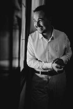 一幅黑白肖像中，一个穿着衬衫的男人微笑着站在室内窗前