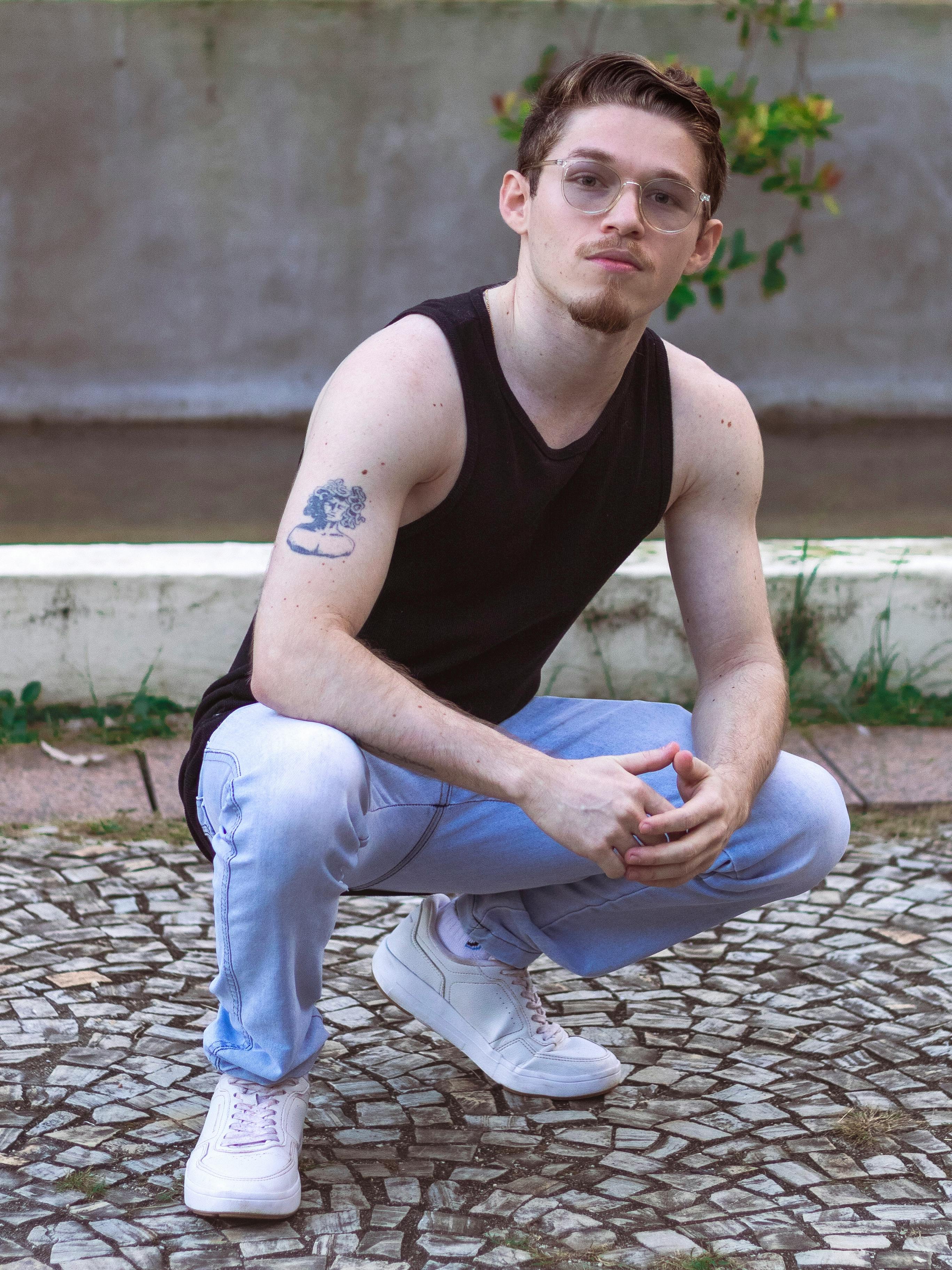 Confident young man in casual attire posing outdoors with visible tattoos.