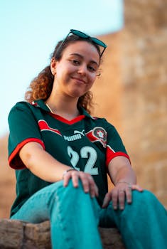 Young woman in Moroccan football jersey sitting outdoors with a soft smile, Rabat, Morocco.