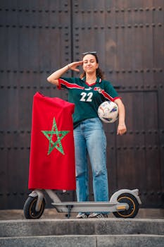 A woman in a football jersey, holding a ball, salutes with a Moroccan flag draped over a scooter.
