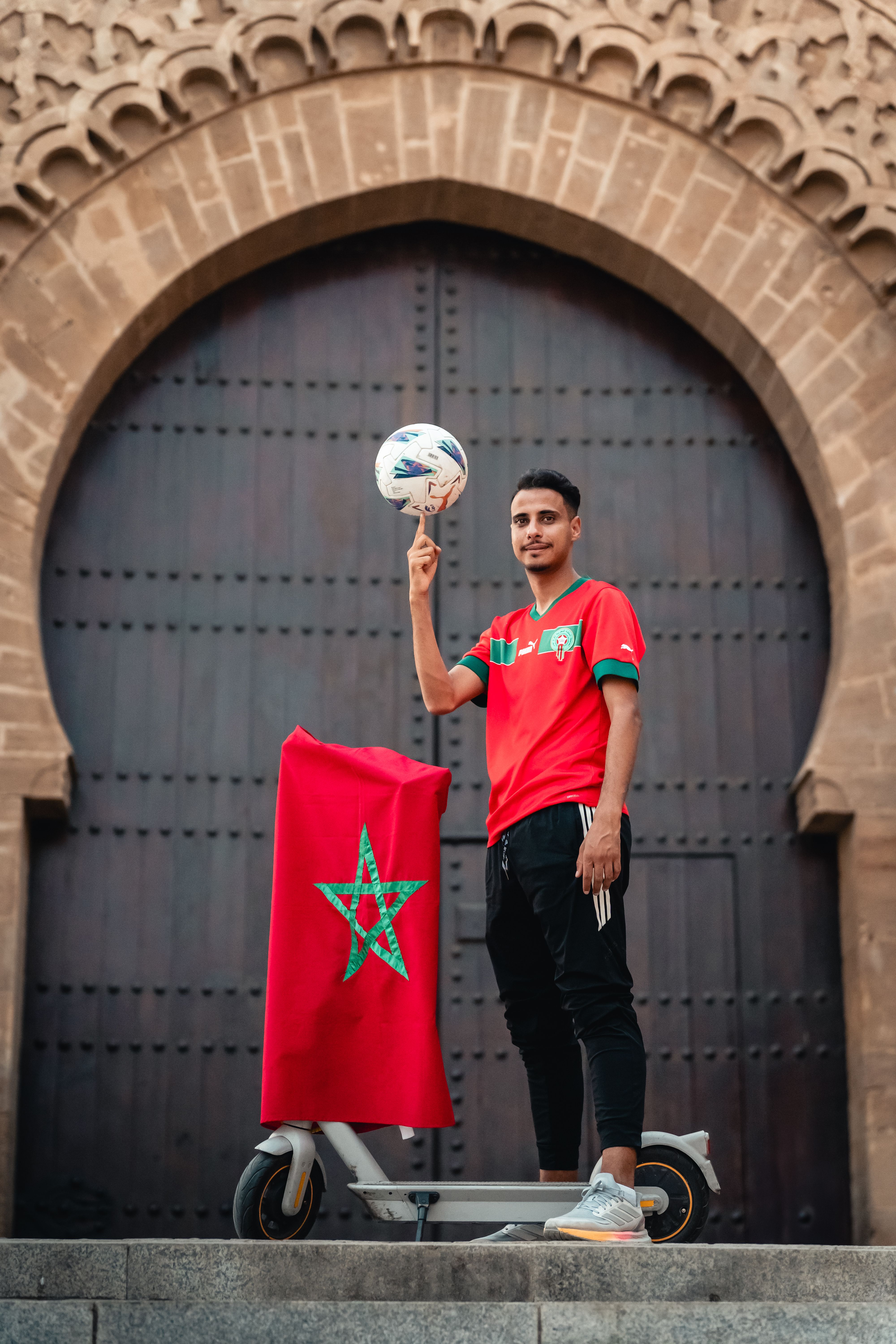 Man in red jersey balances soccer ball atop scooter by Moroccan archway.