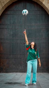 A young woman skillfully balances a soccer ball on her finger against a large wooden door backdrop.