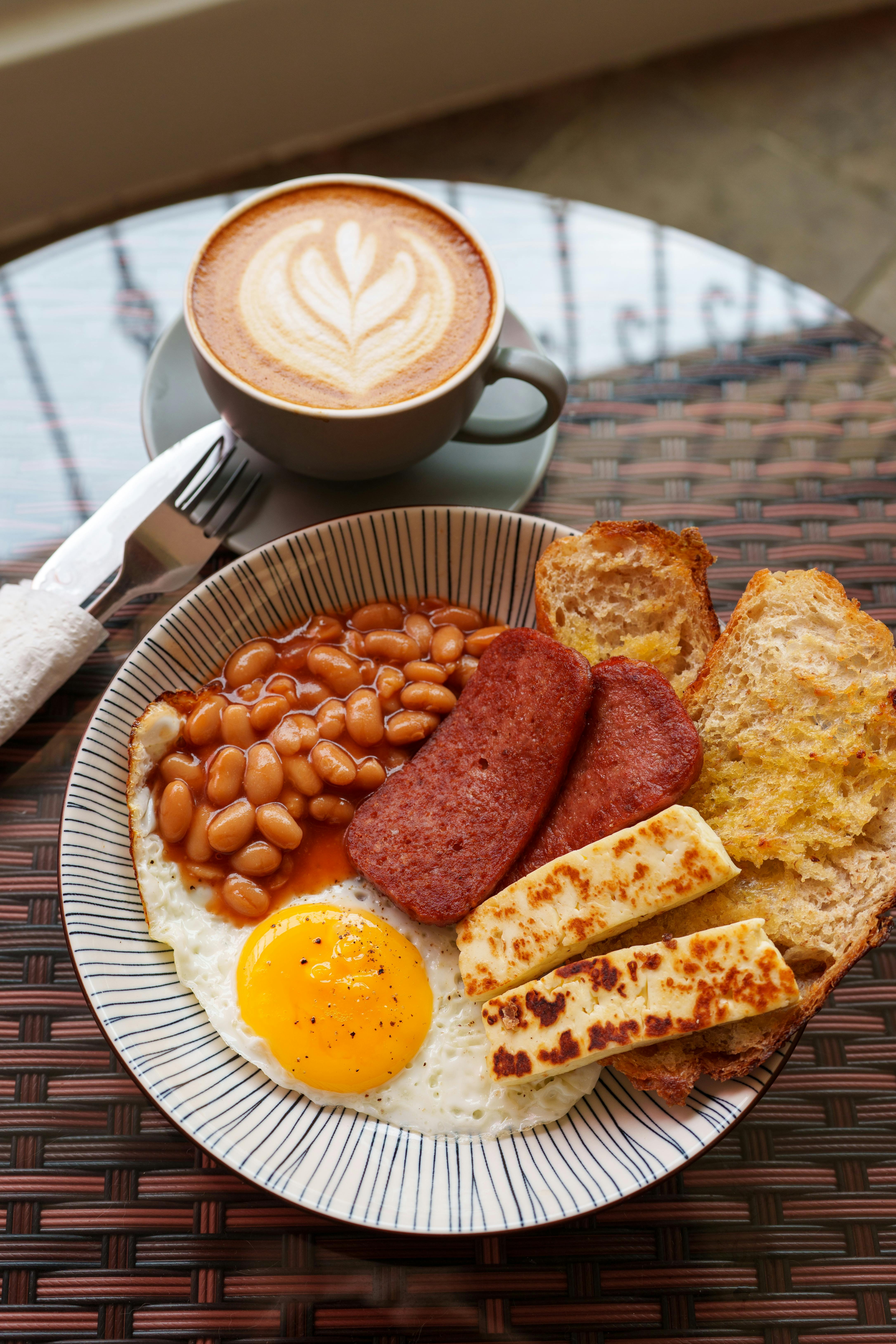 A delicious Full English Breakfast featuring eggs, beans, and toast served with a latte on a glass table.
