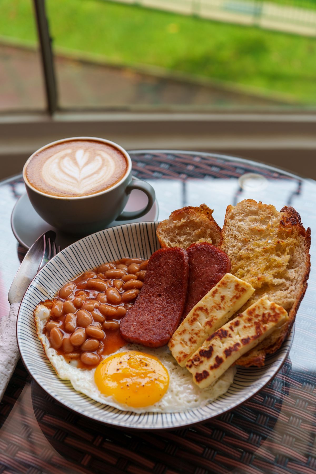 A cooked breakfast with beans, egg and toast beside a coffee, by a window