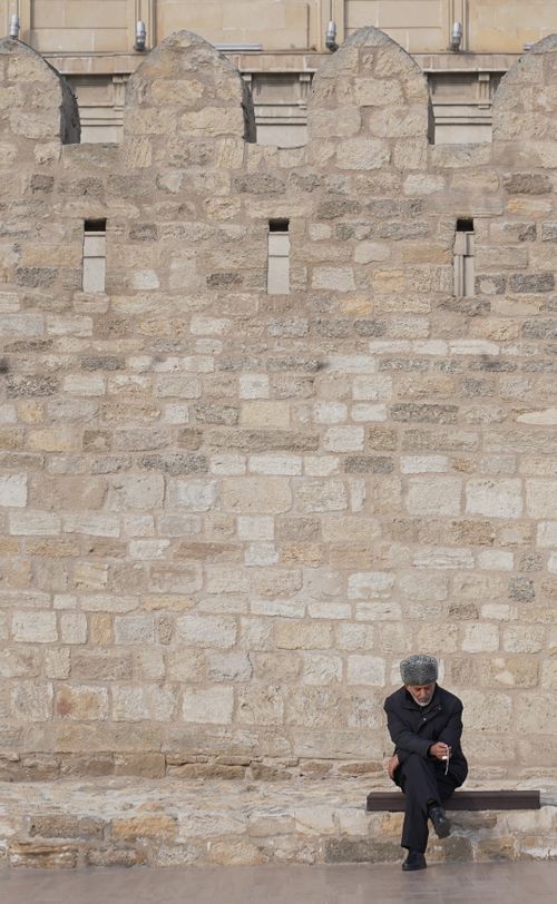 Free Elderly man in fur hat seated near historic stone wall in Baku, Azerbaijan. Stock Photo