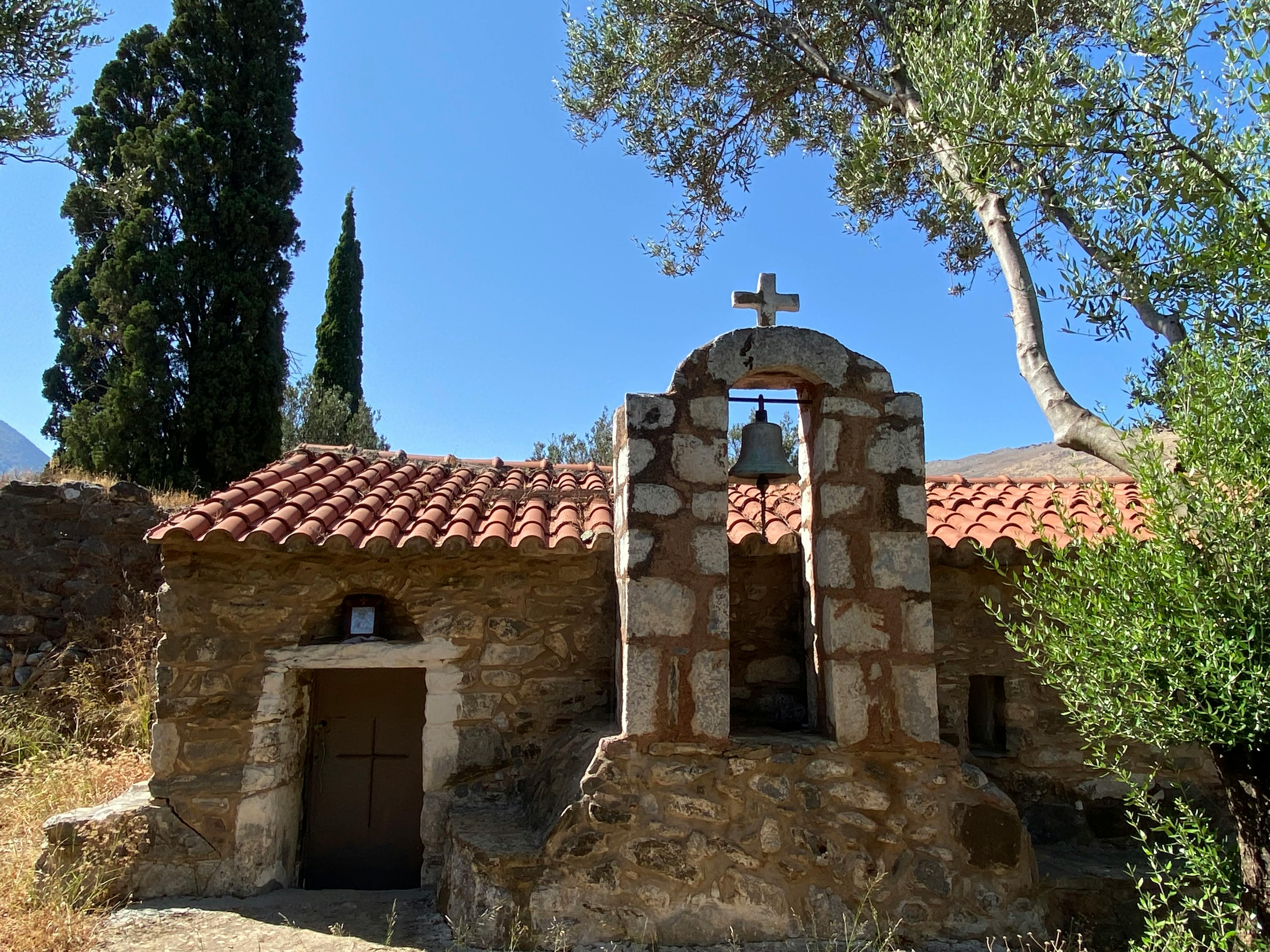 Charming stone chapel with bell tower, surrounded by trees in Greek countryside.