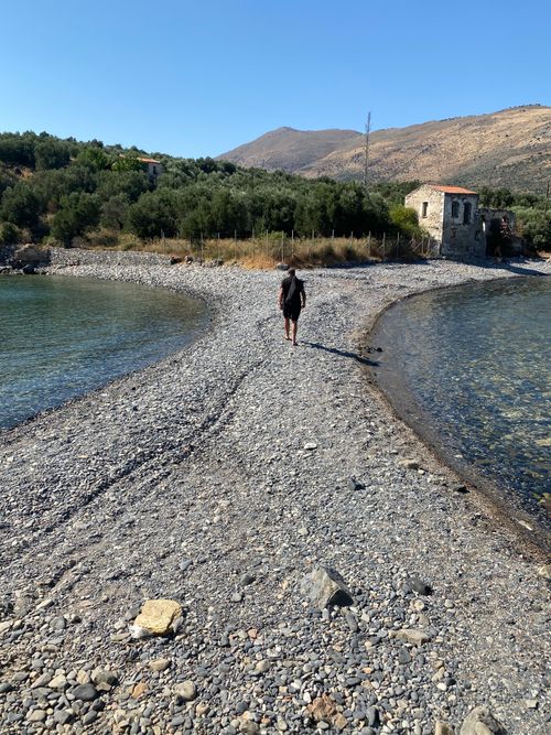 Free A person walks on a secluded pebble beach in Greece with clear waters and an old stone building. Stock Photo