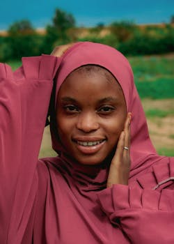 Portrait of a smiling woman wearing a pink hijab, enjoying a sunny day outdoors.