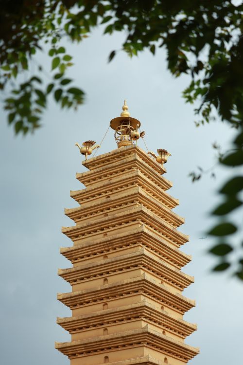 Free A golden pagoda tower stands tall against a cloudy sky, framed by tree leaves. Stock Photo