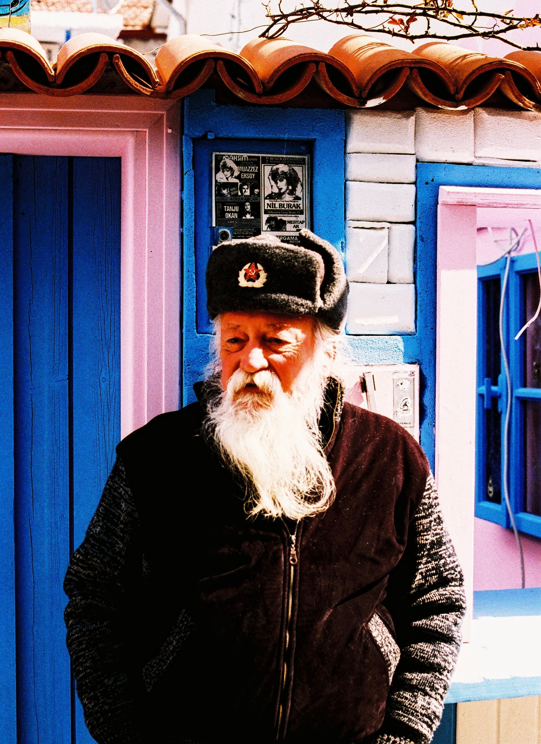 Senior man with a white beard stands thoughtfully against a blue and pink wall in Sığacık, Türkiye.