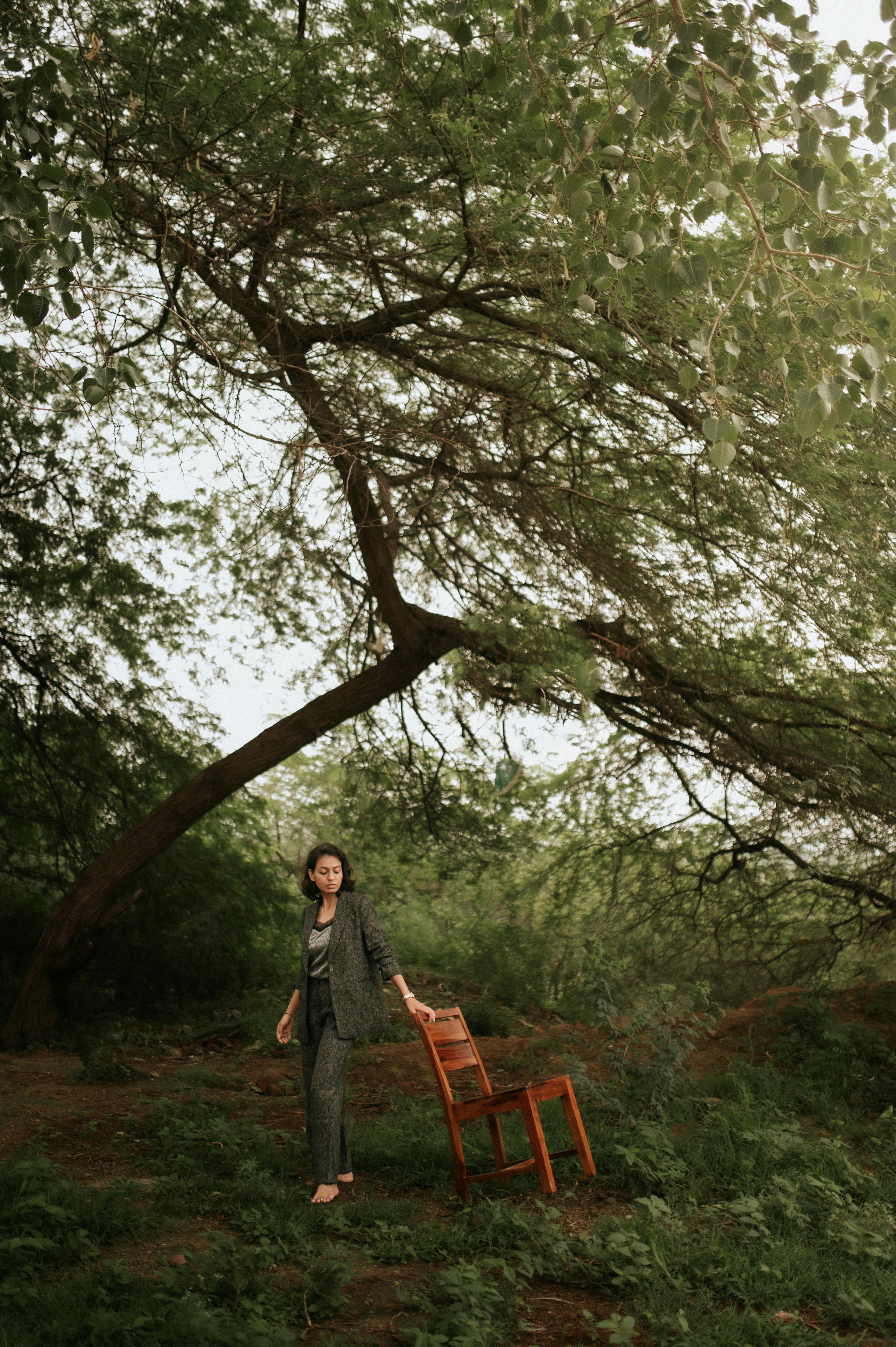 A woman stands by a wooden chair beneath a sprawling tree in an outdoor setting in New Delhi.
