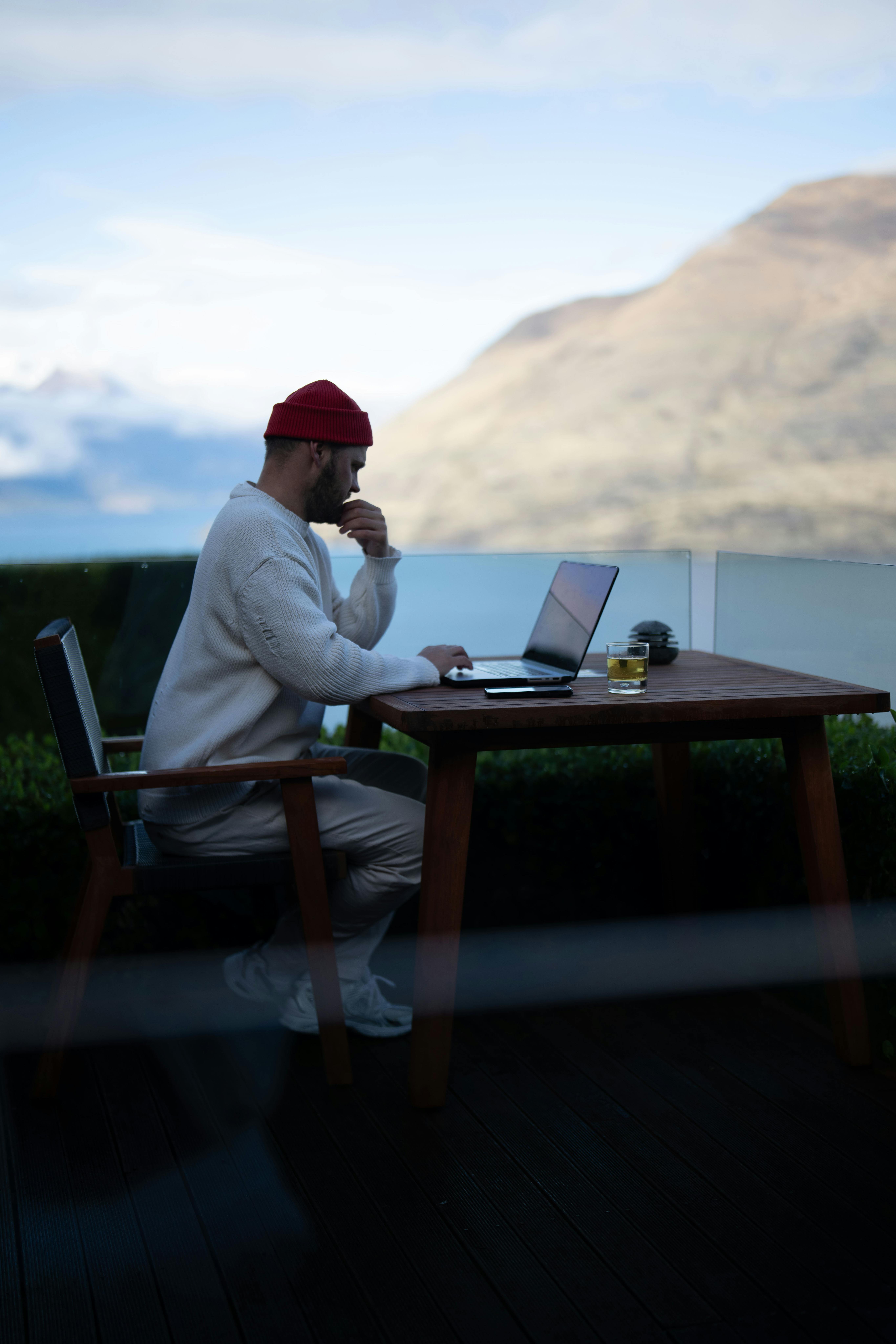 A man in casual attire works on a laptop on a terrace with stunning mountain views in Queenstown, New Zealand.