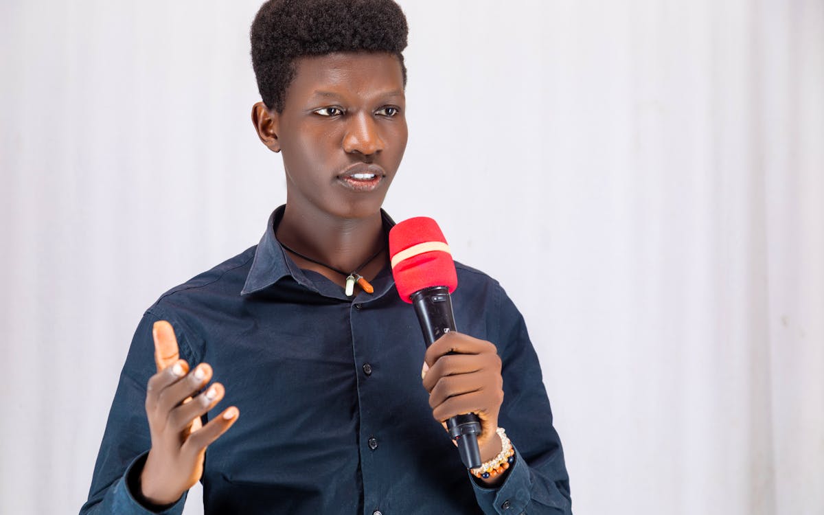 Confident young man presenting with a red microphone, wearing a dark shirt indoors.