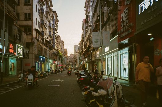 Vibrant urban street scene with people, shops, and scooters in an Asian city at dusk.