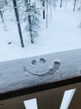 A happy smiley face drawn in snow on a wooden railing in a snowy forest.