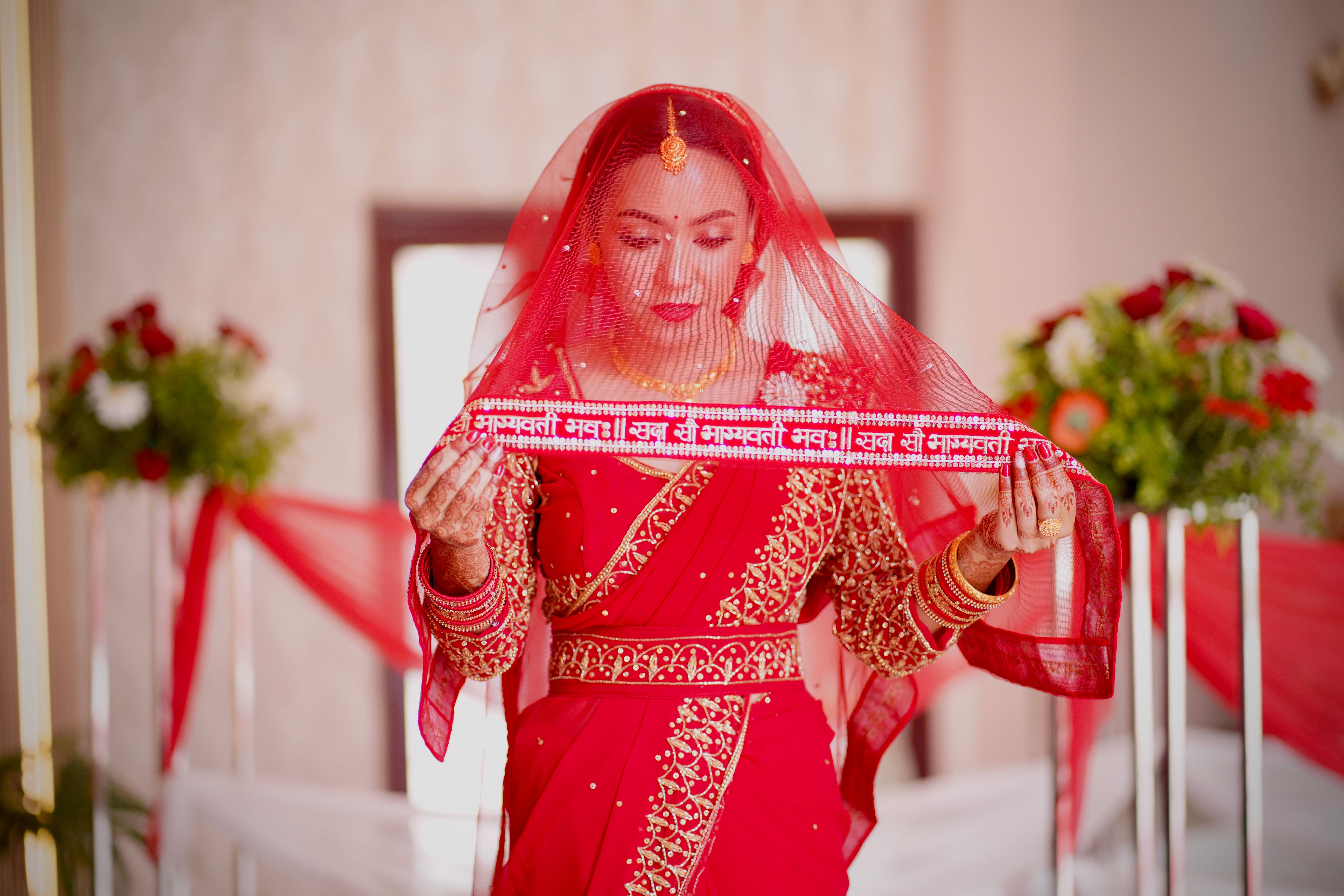 A beautiful Indian bride in red traditional attire, standing indoors with floral decor.