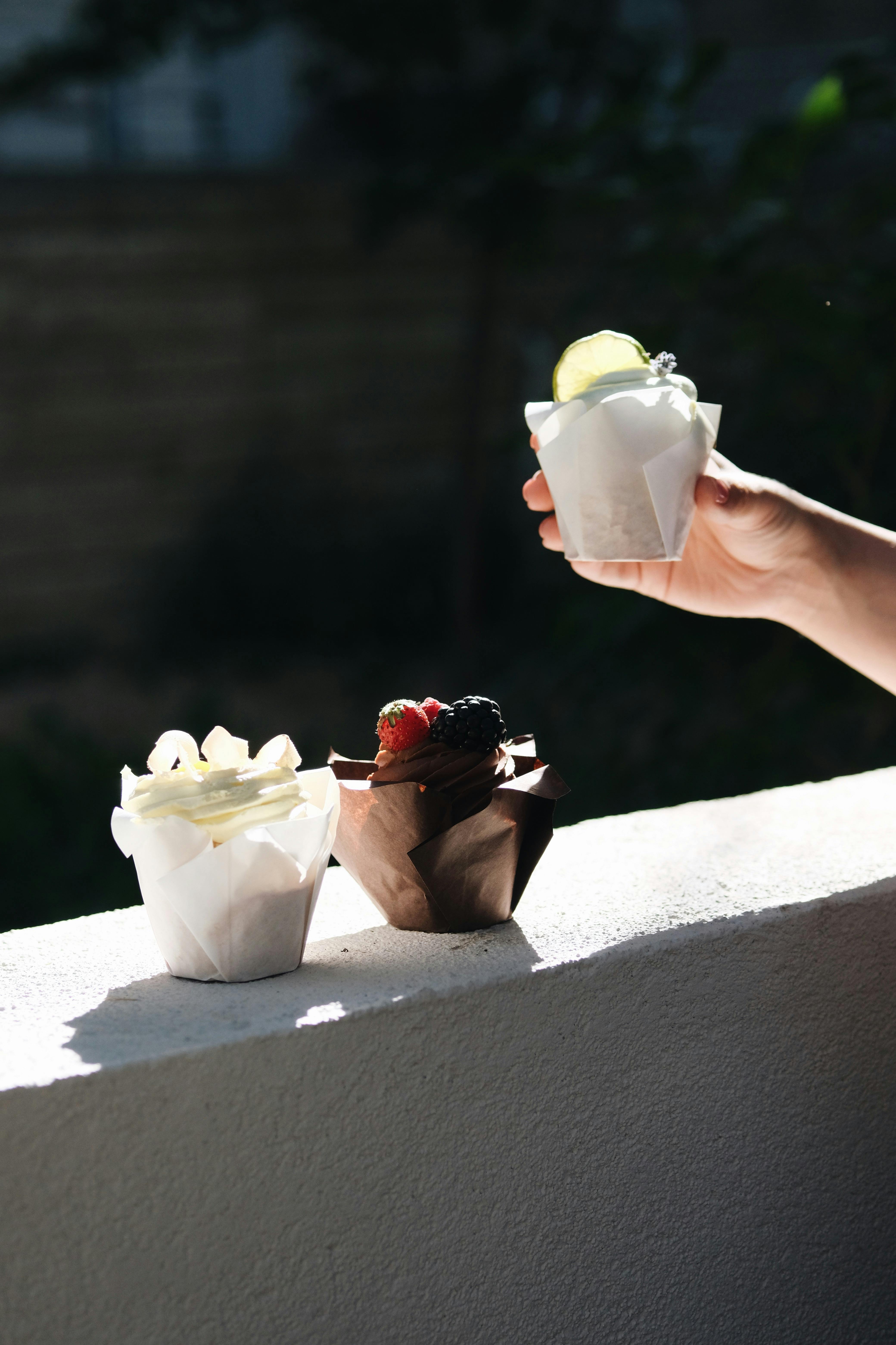 A hand holds a cupcake with a lime slice, next to two on a sunlit ledge.