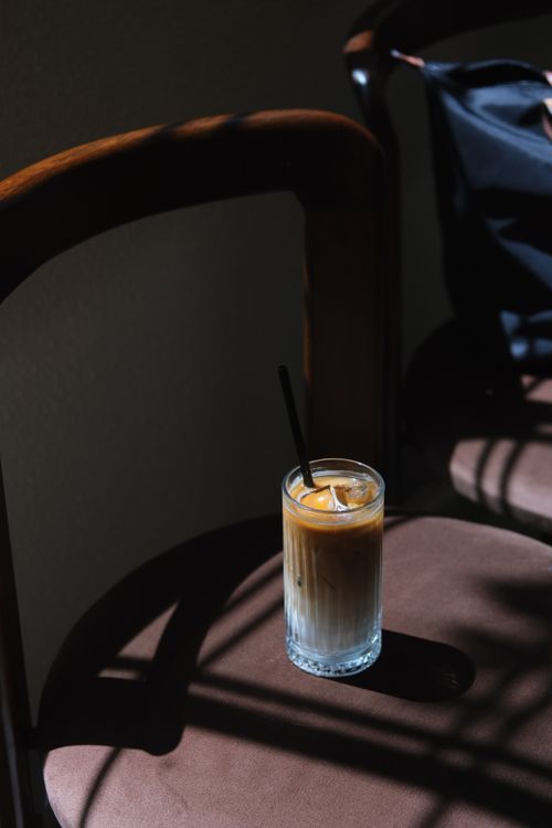 Free A tall glass of iced coffee on a chair, surrounded by dramatic shadows in a sunlit room. Stock Photo