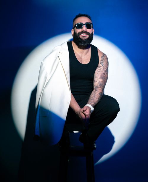 Free Portrait of a stylish man with a tattooed arm, sitting in a spotlight, wearing sunglasses and a white jacket. Stock Photo