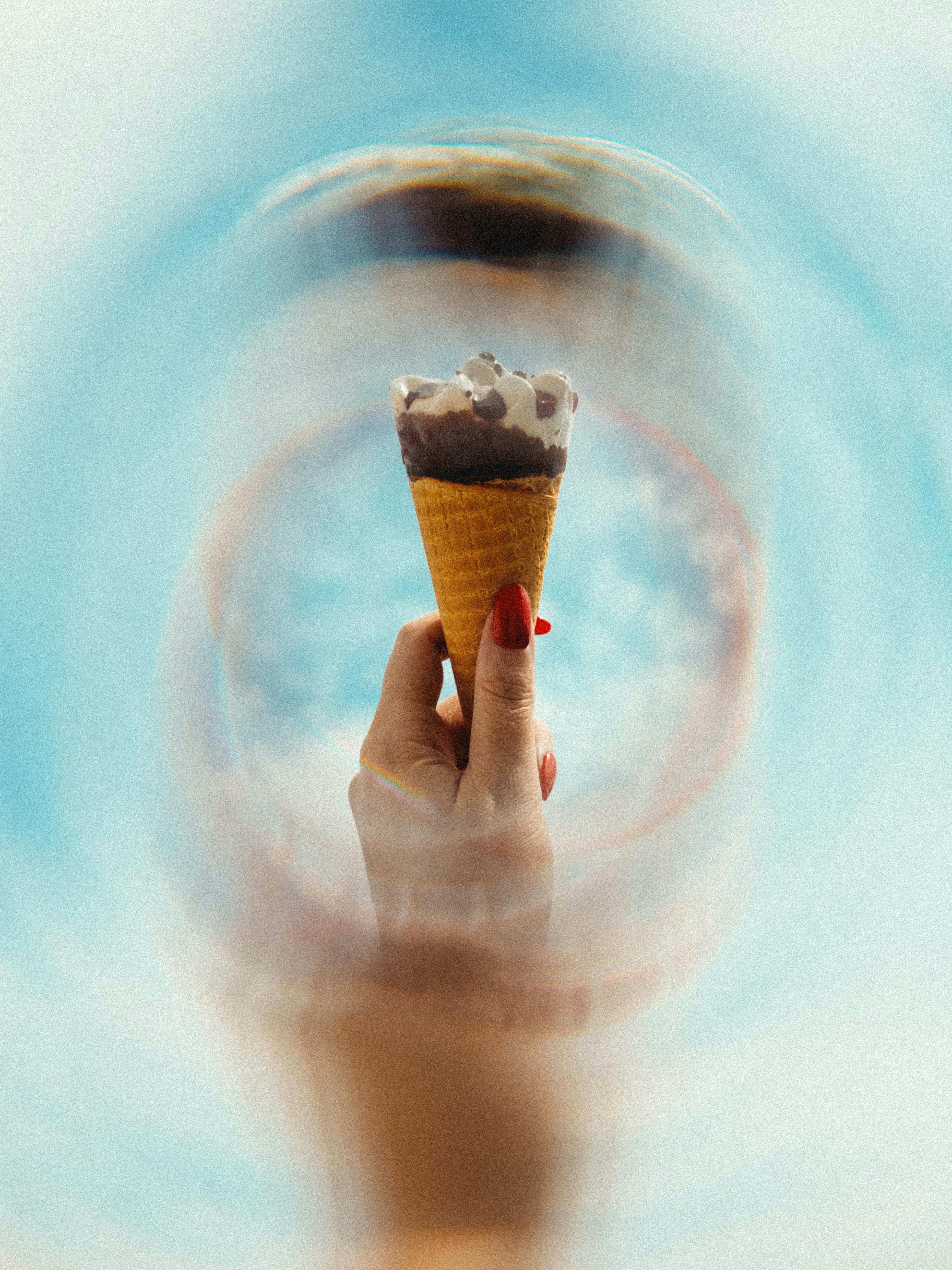 A hand with red nails holding a swirl ice cream cone against a vibrant blue sky, symbolizing summer fun.