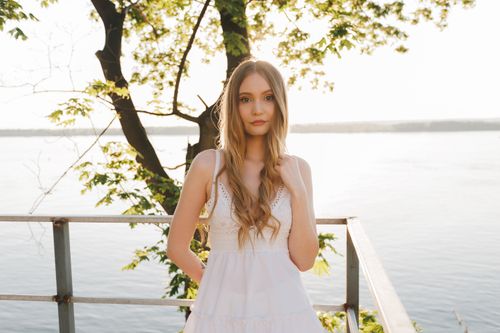 Free A young woman in a white dress stands by the water with sunlight filtering through trees. Stock Photo