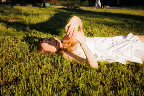Free Young woman in a white dress lying on grass enjoying the warm sunlight outdoors. Stock Photo