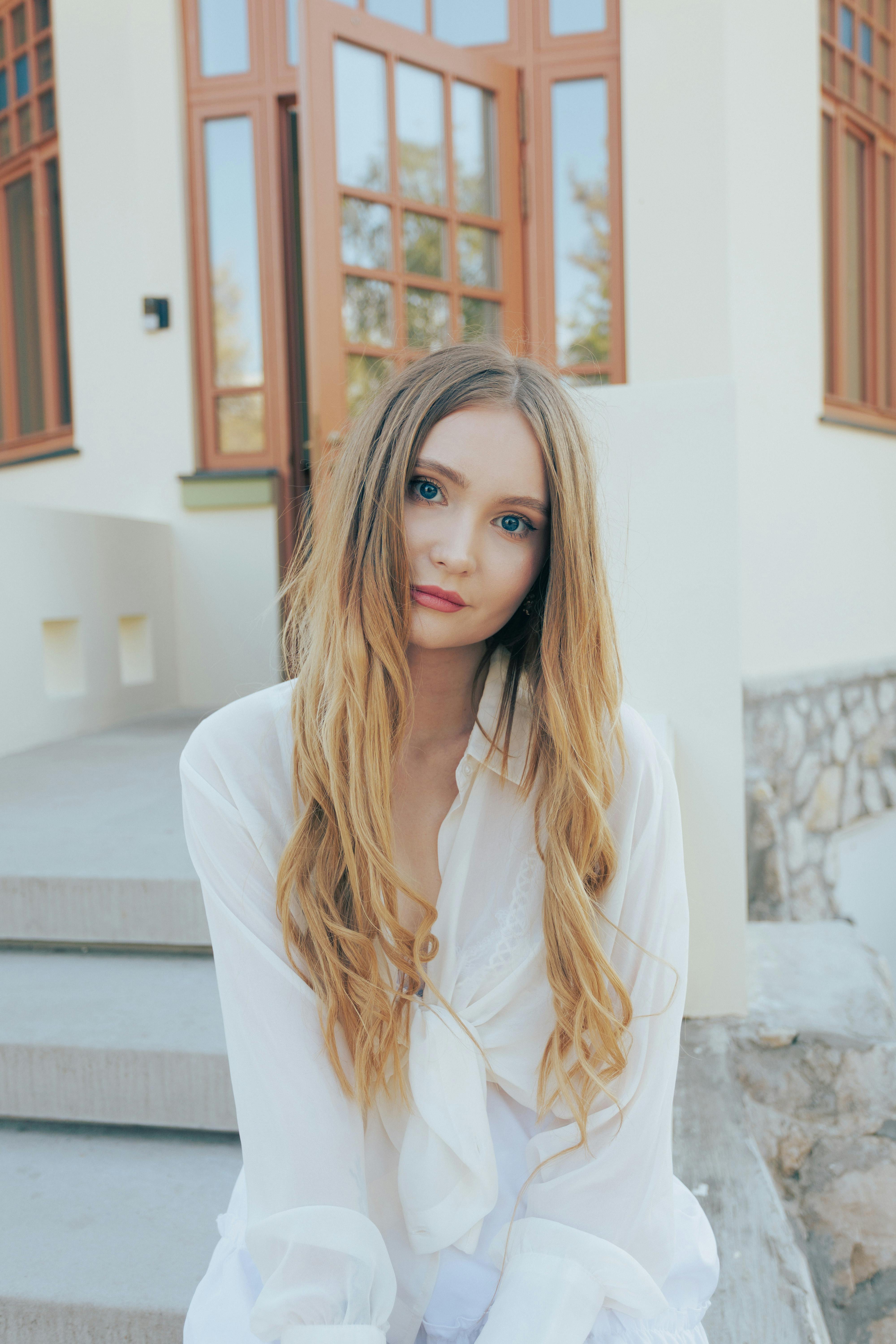 Portrait of a young woman with long hair seated on outdoor steps, wearing a white blouse.