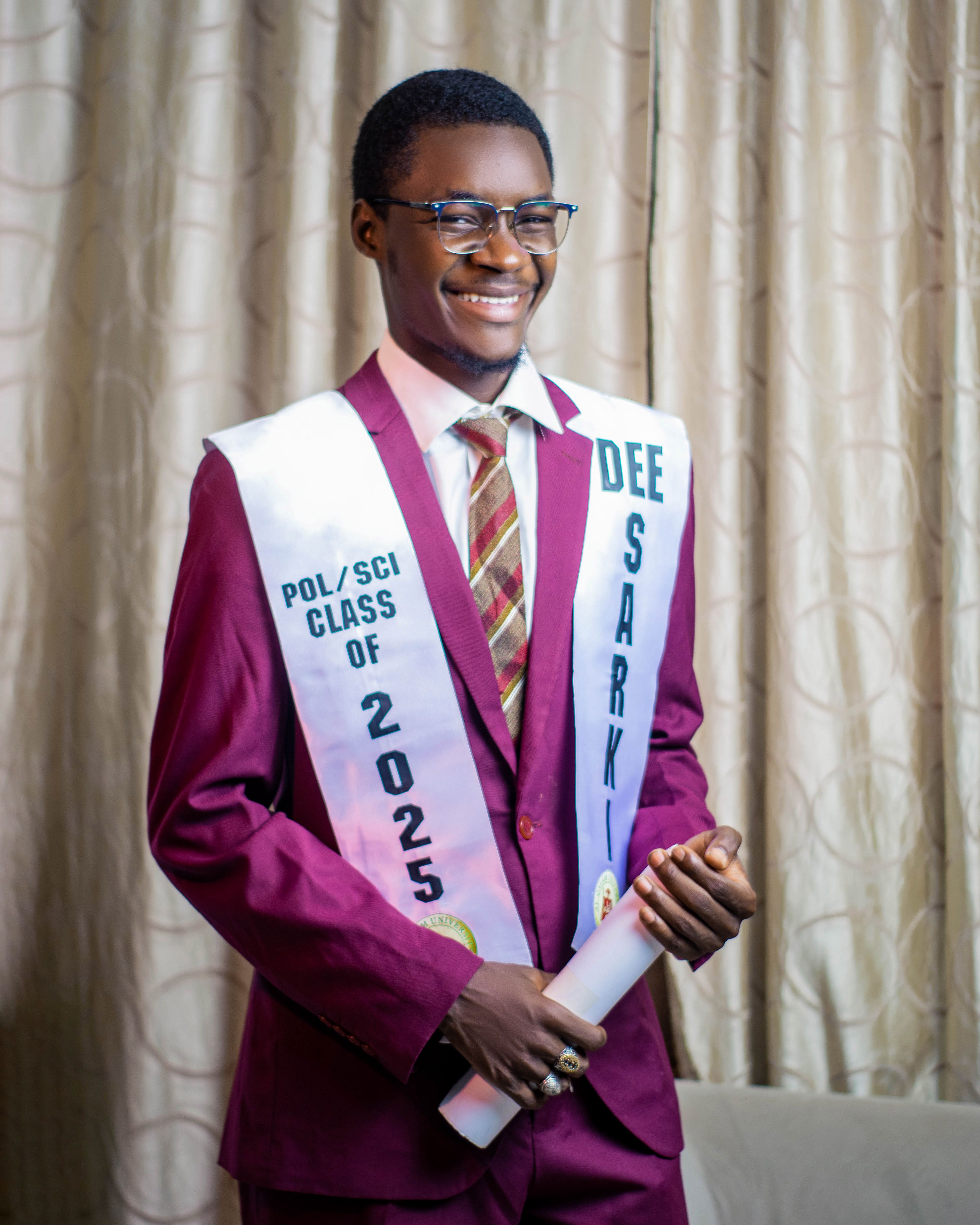 Young graduate smiling in maroon suit with sashes, holding diploma - Political Science Class of 2025.