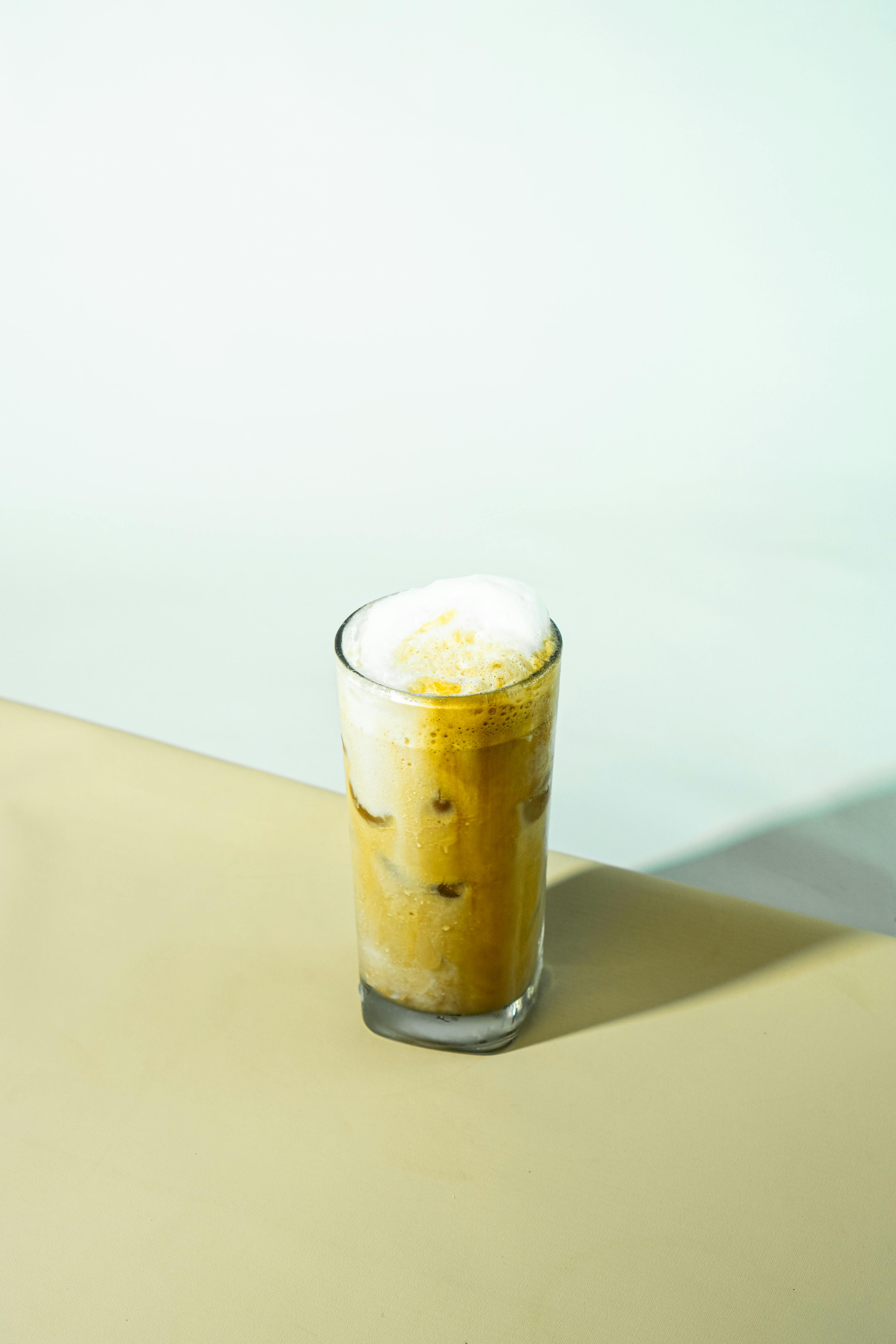 Tall glass of iced coffee with creamy foam on a neutral background.