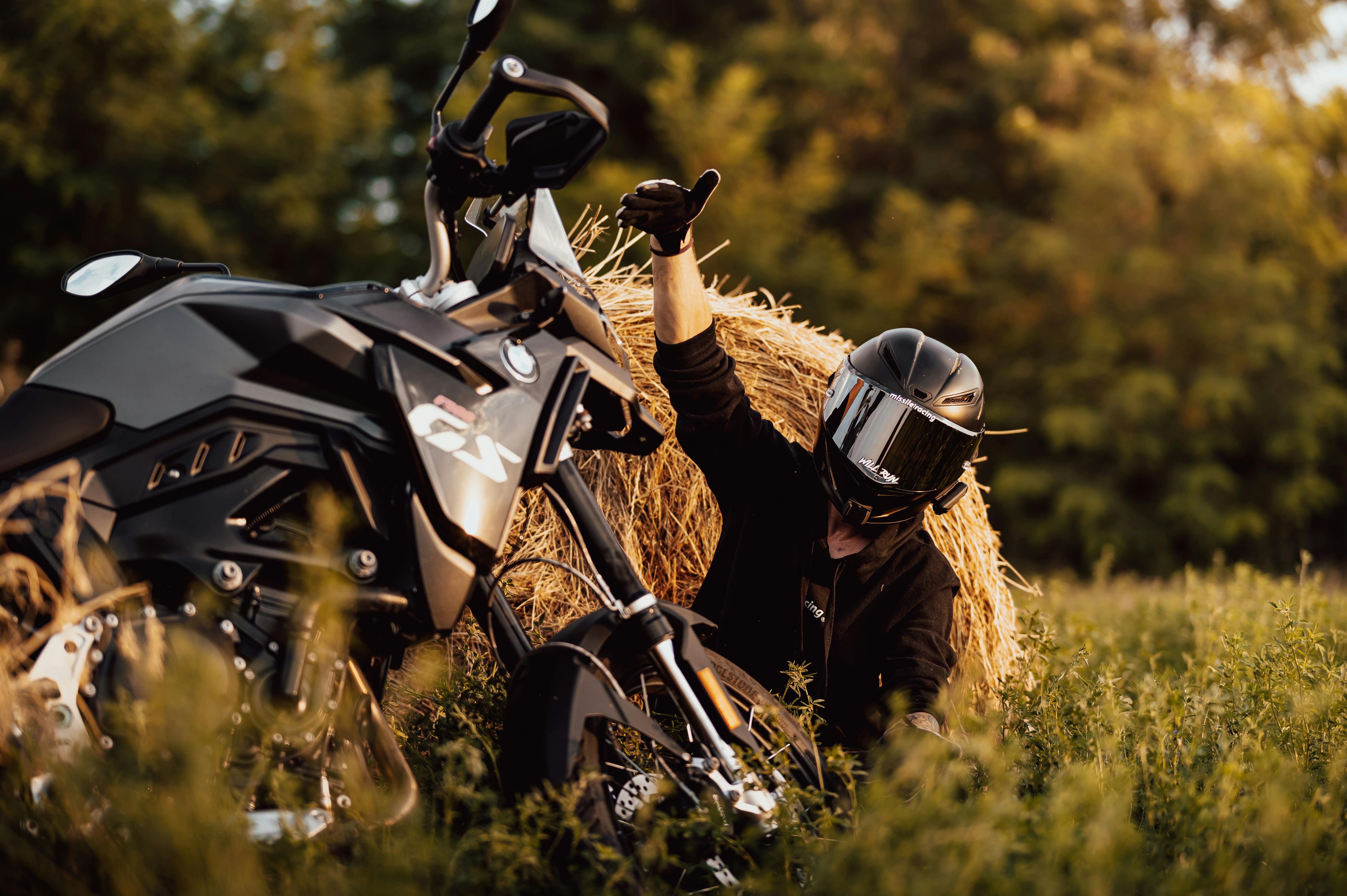 Un motard lève les pouces, se reposant contre une botte de foin avec une moto dans un champ au coucher du soleil.