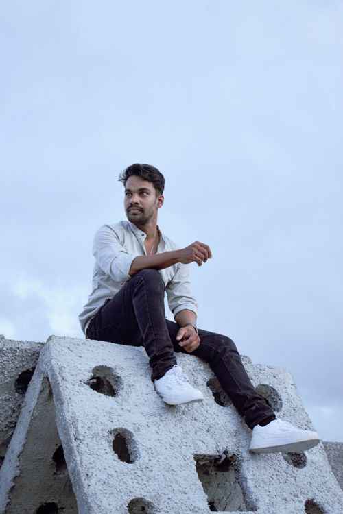 Free Young man in casual attire sitting on a concrete block with a cloudy sky backdrop. Stock Photo