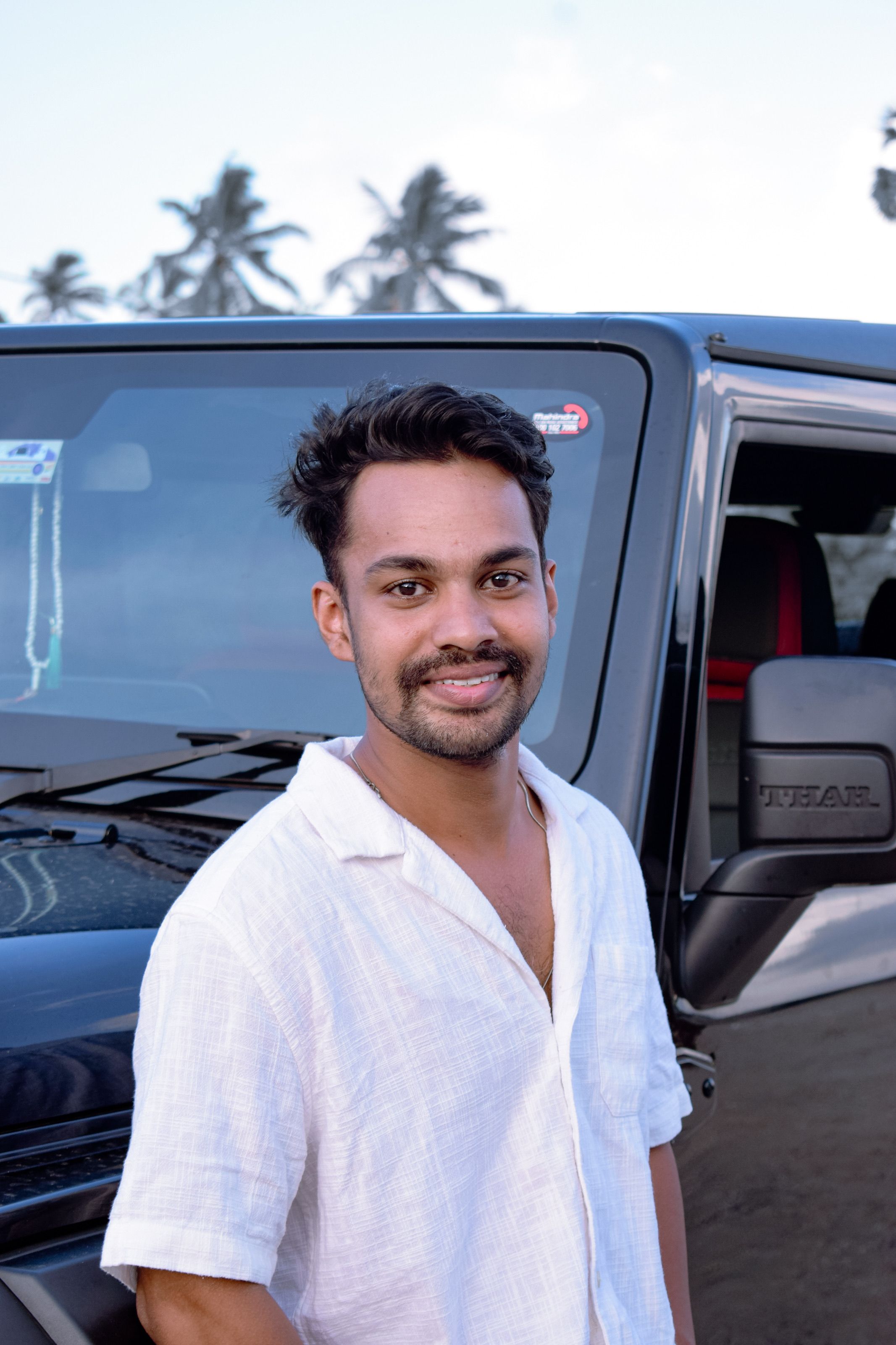 Smiling man in a white shirt standing next to an SUV with palm trees in the background.