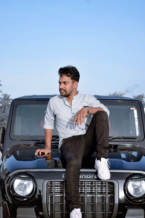 Free A stylish man in casual attire sits on an SUV's hood against a clear blue sky. Stock Photo