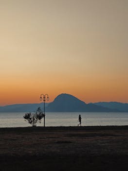A solitary figure walks along the shoreline at sunset, with a mountain in the distance.